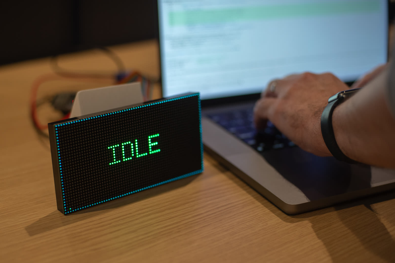ClaudePanel on a desk beside a laptop: a 64×32 LED matrix showing IDLE in green dot-matrix letters, ringed by a cyan border, while someone types on the laptop behind it.